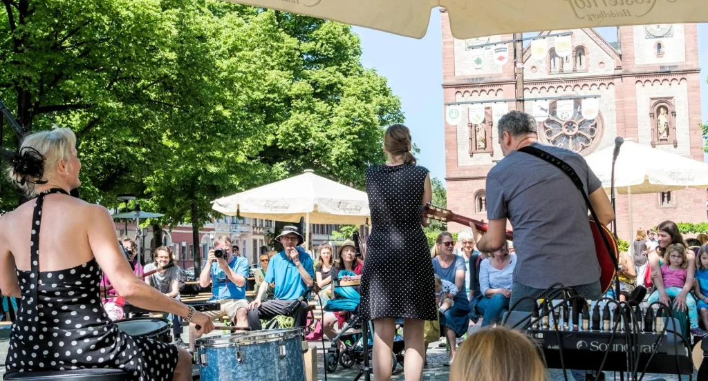 Foto von Valentin Bachem. Rückseite der Band. Dahinter ist das sitzende und lauschende Publikum. Ein Fotograf fokusiert auf die Frau an den Drumms.
