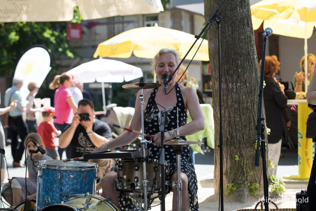 Foto von Rainer Herbold. Die Sängerin sitzt an den Drumms und singt ins Mikrofon. Dahinter eine Person, die gerade ein Foto macht.
