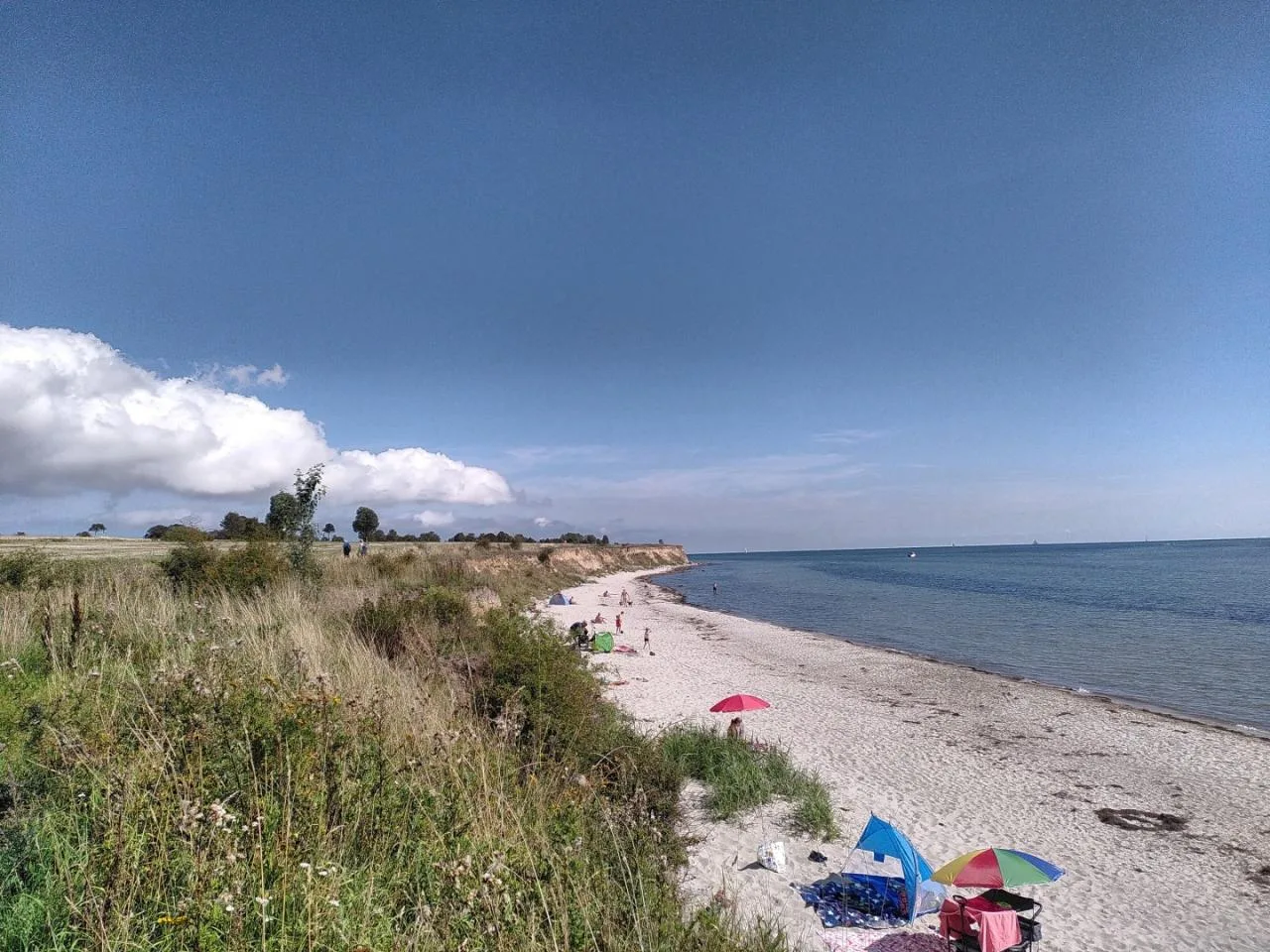 Strandfoto von Fehmarn. Es gibt einige Gäste am Strand. Der Himmel und das Meer sind blau. Im Hintergrund Wolken und die Steilküste.