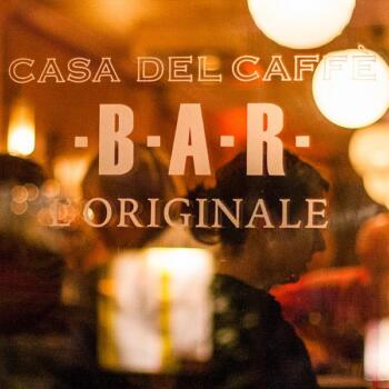 Blick von außen ins Café, das Innere ist unscharf, der Schriftzug mit dem Name des Cafés ist scharf abgebildet: "Casa del Caffé BAR Originale"