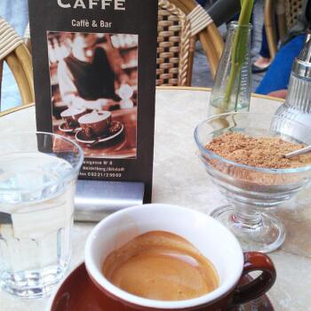 Eine Tasse mit frischem Espresso mit schöner Crema, einem Glas Wasser und loser brauner Zucker in einem Schälchen stehen auf einem runden Tisch