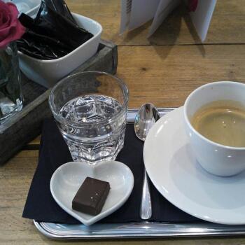 Tasse doppelter Espresso, serviert auf einem silbernen Tablett mit Schokoladenpraline. Links davon steht eine rote Rose in einem Glas.