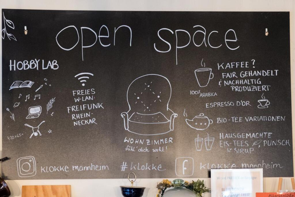 Eine schwarze Tafel mit der Aufschrift Open Space. Unter einem Stuhl steht "Wohnzimmer fühl dich wohl!". Es gibt weitere Infos zu den Getränken und Social Media.