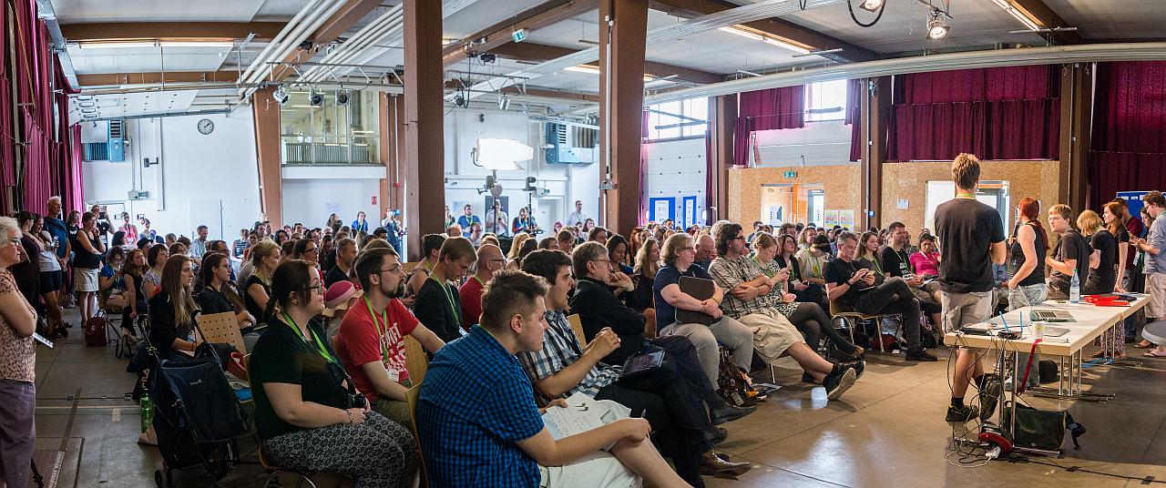 Panorama von der Begrüßung. Viele Menschen sitzen in einem großen Saal und werden vom Orateam begrüßt