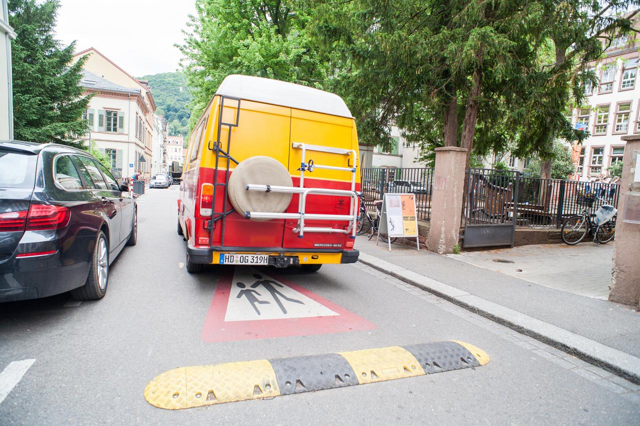 Theaterstraße und ein bunter Bus. Auf dem Boden ist ein Vorsicht-Schild mit spielenden Kindern aufgemalt und eine Bodenschwelle.