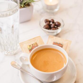 Eine Tasse Espresso, ein Glas Wasser, Blumen und ein Schälchen mit einer Schokoladensüßigkeit stehen auf einem Tisch