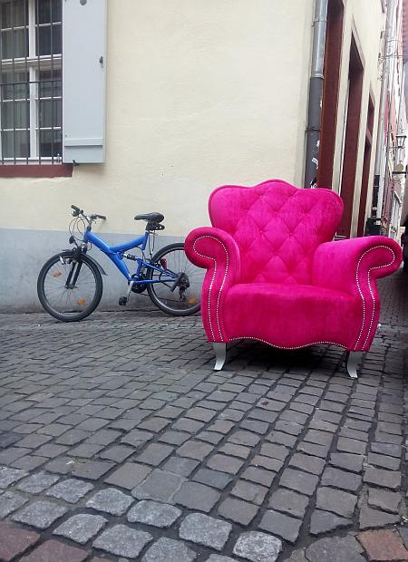 Ein schicker Sessel in grellem Rosa steht auf dem Kopfsteinpflaster