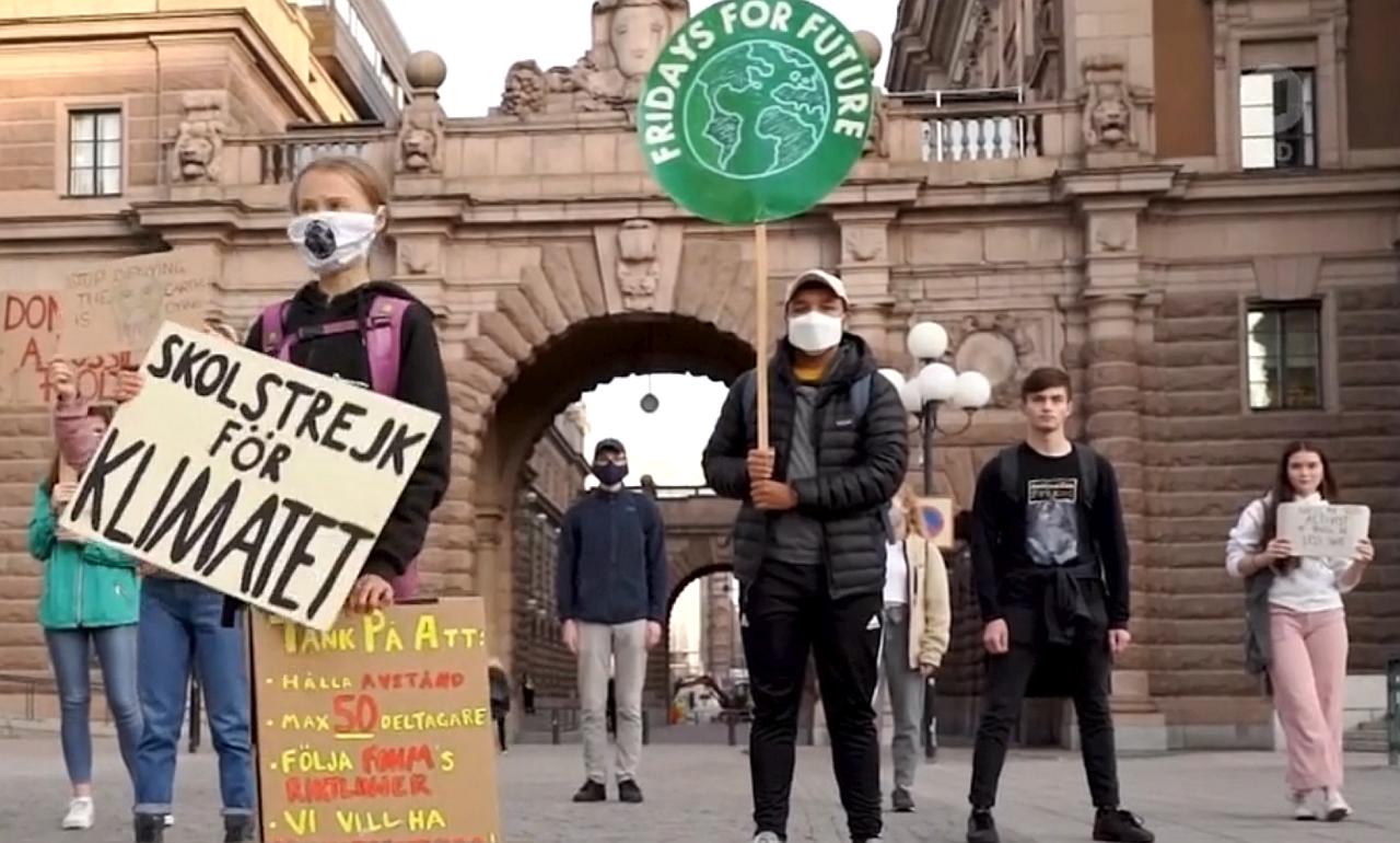 Bildschirmfoto aus dem Trailer. Klimaaktivisten stehen mit Maske und Plakaten vor einem Gebäude