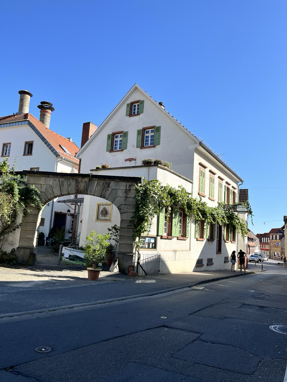 Ein Gebäude mit einem Torbogen und grünem Bewuchs. Es liegt an einer Straße, die den Berg herauf führt. Der Name ist Bachlenz. 