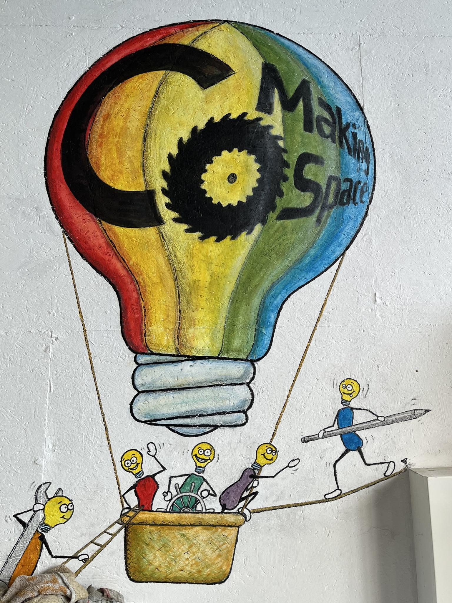 Zeichnung eines regenbogenfarbenen Ballons in Form einer Glühbirne. Im Korb drei kleine Glühbirnen. Eines davon am Steuerrad. Zwei weitere sind dabei zuzusteigen. Das eine trägt einen Schraubenschlüssel und das andere einen Bleistift. Die im Korb helfen beim Zustieg. 