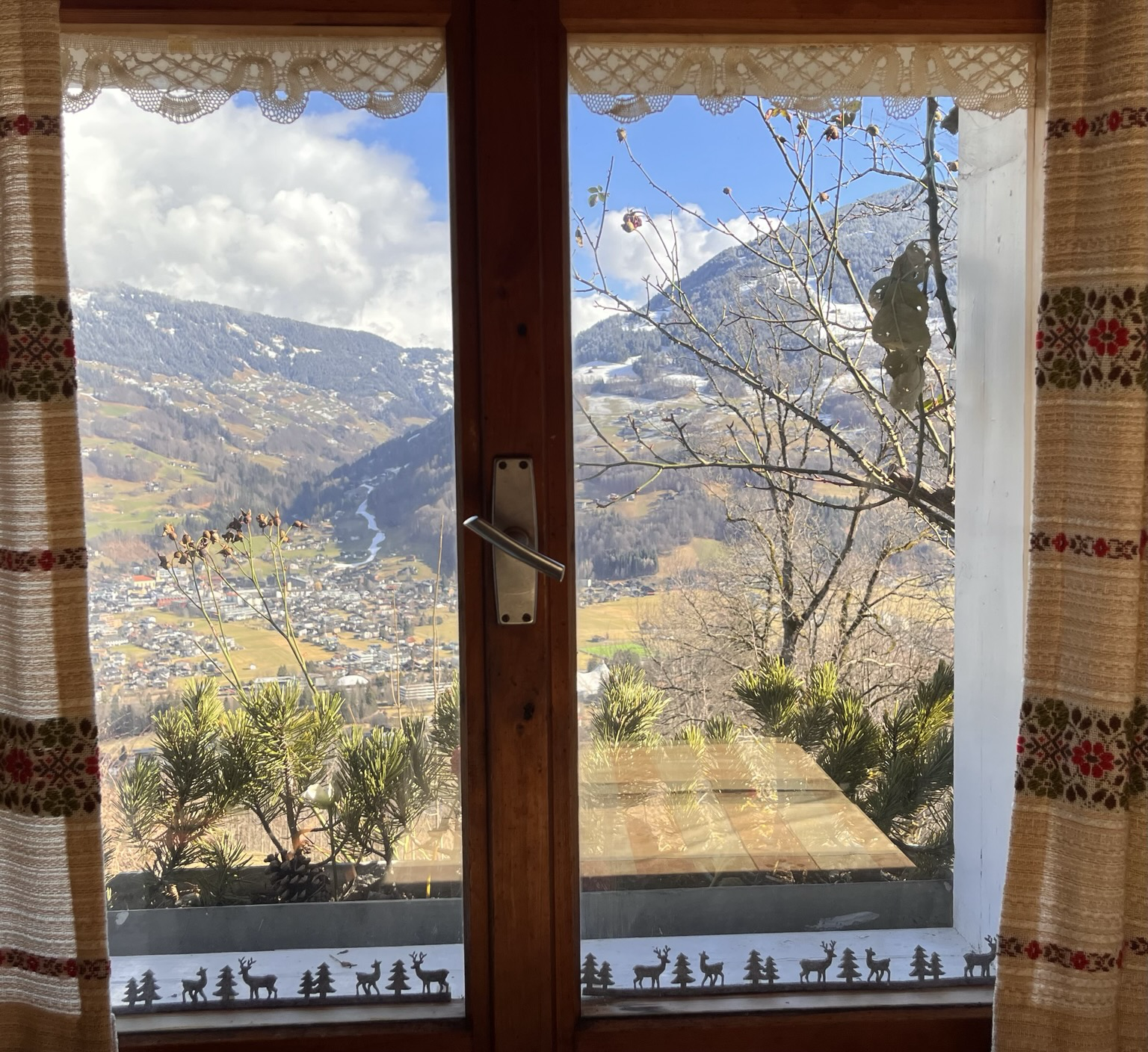 Fenster in einer Hütte mit Blick nach draußen. Es zeichnet sich eine Berglandschaft der Alpen ab. Blick runter ins Tal auf einen Ort. Auf den Bergen liegt Schnee 