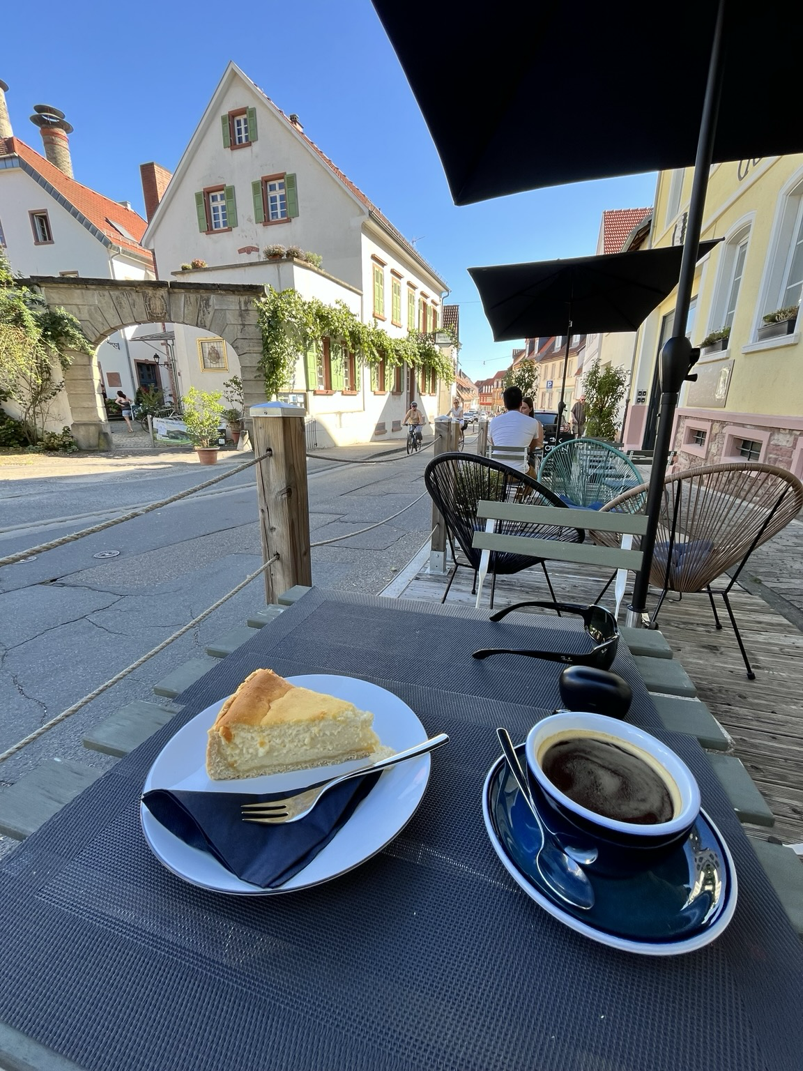 Weitwinkelaufname der gleichen Situation. Es ist eine Terrasse mit Stühlen und Tischen zu erkennen. Auf dem mir nächsten Tisch steht eine Tasse Kaffee und ein Teller mit einem Stück Käsekuchen. 