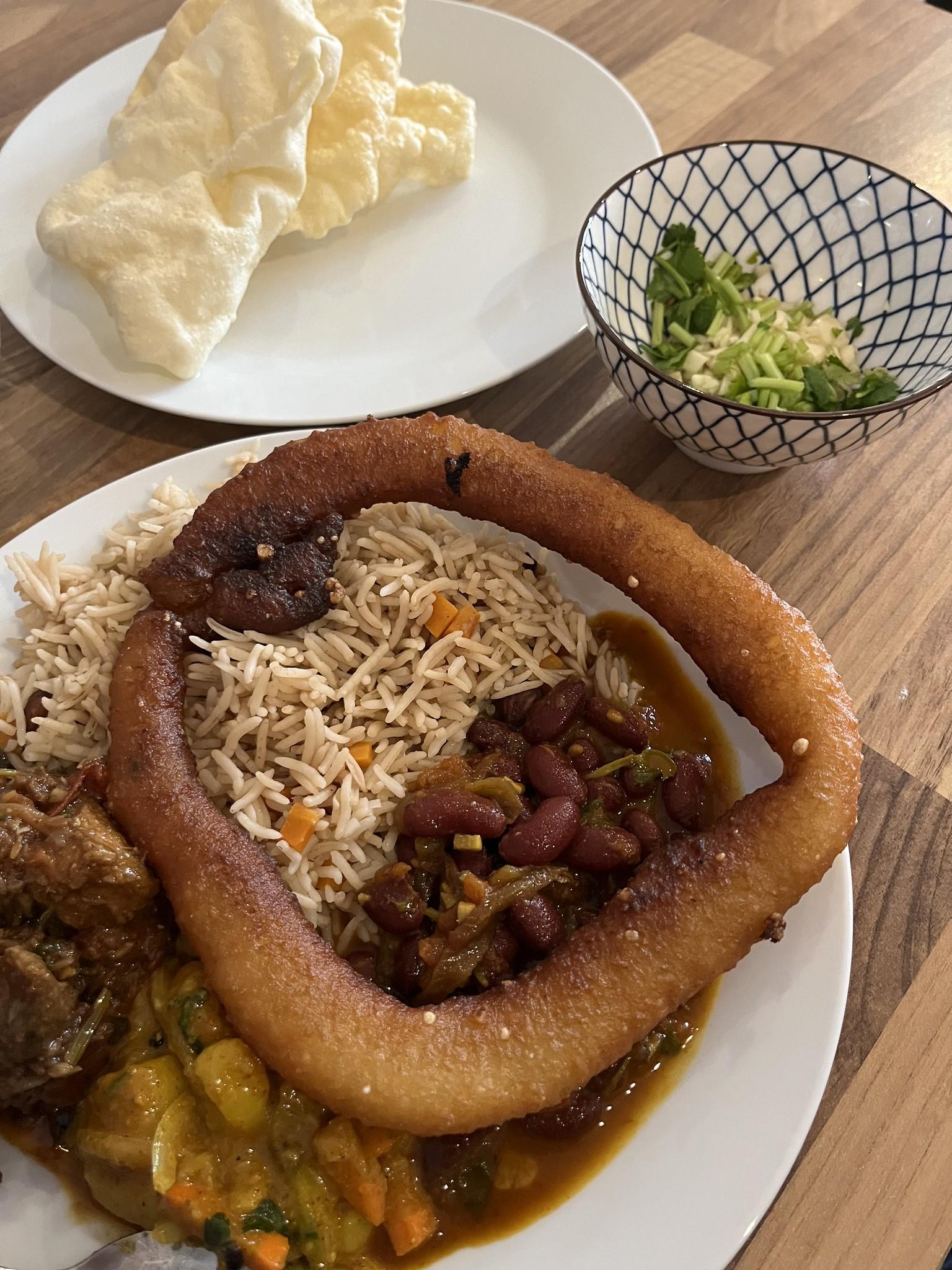 Ein Teller mit nepalesischem Essen. Reis, Gemüse, Fleisch, Soße und Bohnen. Darauf liegt ein Teiggebäck. Dahinter in einer Schale Koreander, Knoblauch und Papadam.